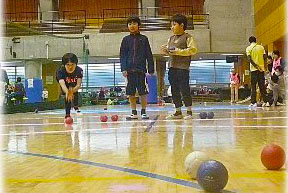 体育館で3人の男子が手のひら大のボールを転がす競技をしている写真