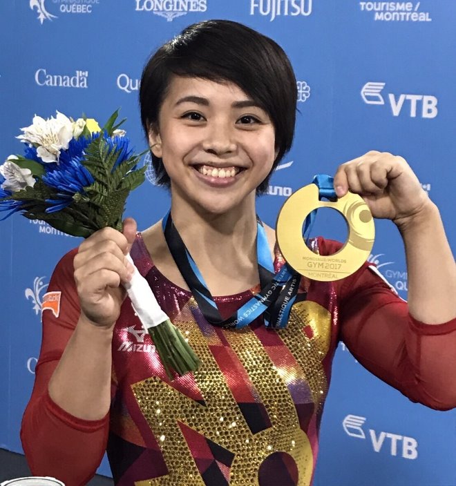 赤いレオタードを着た短い髪の女性体操選手が右手に花束、左手に金メダルを持って笑顔で写っている写真