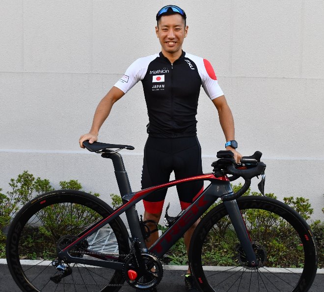 日本代表ユニフォームを着た男性がバイクを持って立っている写真