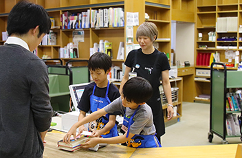エプロンをつけた小学生が図書館のカウンターで本の貸出し体験をしている写真