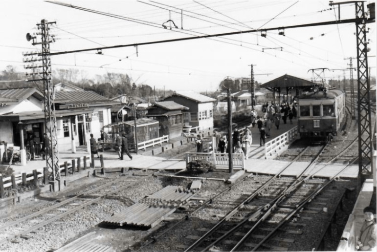 ホームに停車している一両編成の電車と少し離れた場所に建つ駅舎、その間を歩く人々の白黒写真